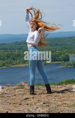 Beautiful red head in skinny jeans, black boots, and white top, on mountain top with wind in hair and a lake below. Stock Photo