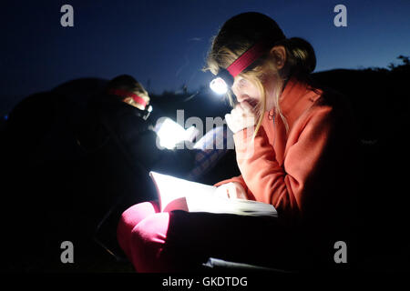 A girl reads a book, and sucks her thumb, by torchlight whilst camping under a clear sky Stock Photo