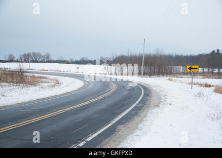 A two lane blacktop road next to a snow covered field curves past an arrow sign on a gray winter day Stock Photo