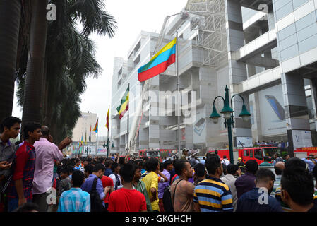 Bangladeshis gather to watch rescue operations outside of the Bashundhara City shopping mall that caught fire in Dhaka, Bangladesh. On August 21, 2016 Fourteen firefighting units are working to control the flame. The fire broke out at shop 360 on block C of the mall which is a shoe shop. The fire broke out at around 11:25am and suddenly spread on the floor, said Abdur Rahman, duty officer at fire service control room. Stock Photo