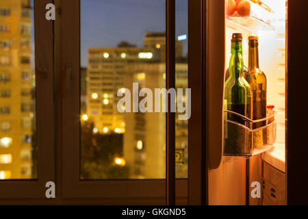 Open door of refrigerator with wine bottles and view of city through home window in night Stock Photo