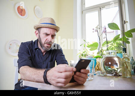 Hipster with hat and beard sitting at home in kitchen with mobile phone Stock Photo