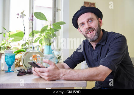 Hipster with beard and hat looking to camera sitting on a table in his ktichen Stock Photo