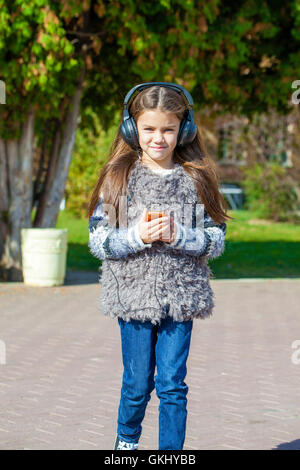 Beautiful little girl listening to music on headphones in autumn park Stock Photo