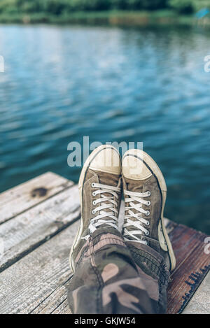 Man with crossed legs relaxing on riverbank pier, casual young guy wearing sneakers sitting by the river in summer afternoon Stock Photo