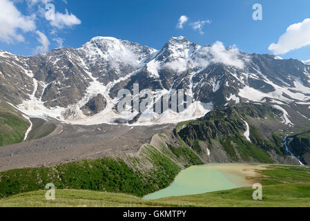 The bluish-green mountain lake against a panorama of snow-covered mountains and peaks with the glaciers Stock Photo