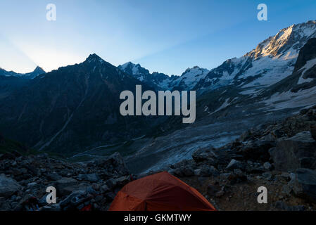 Panorama of snow-covered mountains and peaks with the glaciers which are flowing down to the valley with orange tent among stone Stock Photo
