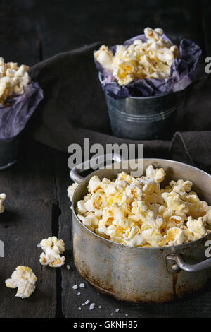 Prepared salted popcorn served with sea salt in small buckets and vintage aluminum pan on old wooden kitchen table. Dark rustic  Stock Photo