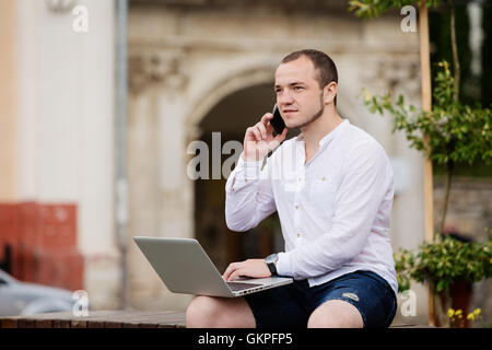 young man on stone bench with laptop and the phone Stock Photo