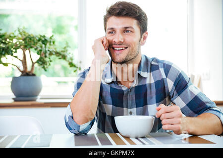 Happy young man in checkered shirt using smartphone and having breakfast on the kitchen Stock Photo