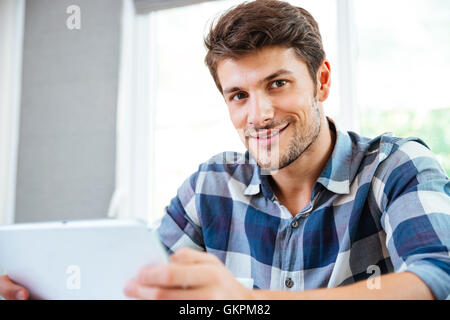 Portrait of happy young man in checkered shirt using tablet at home Stock Photo