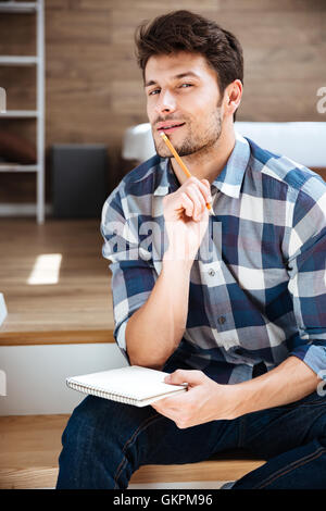 Thoughtful young man in checkered shirt with notepad and pencil sitting and thinking at home Stock Photo