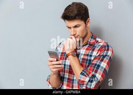 Pensive young man in checkered shirt using mobile phone over grey background Stock Photo