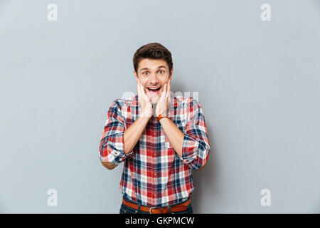 Surprised excited young man in plaid shirt standing with mouth opened over grey background Stock Photo