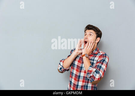 Scared shocked young man in plaid shirt looking away and shouting over grey background Stock Photo