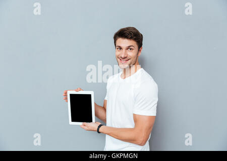 Smiling handsome young man standing and holding blank screen tablet over grey background