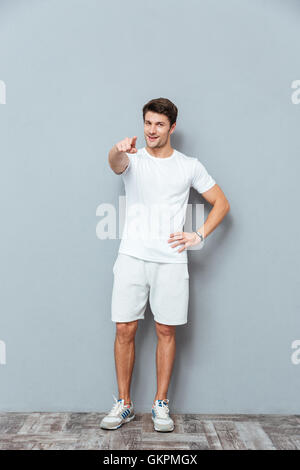 Full length of handsome smiling young man standing and pointing on you over grey background Stock Photo