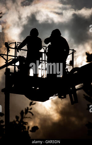 Two London firefighters silhouetted against sun and smoke whilst operating a height platform at the scene of a fire in London Stock Photo