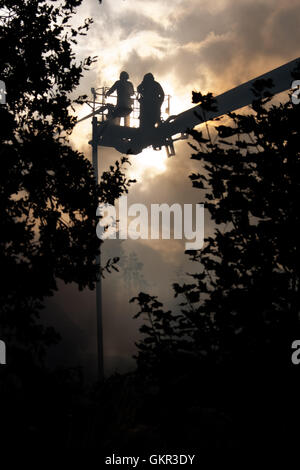 Two London firefighters silhouetted against sun and smoke whilst operating a height platform at the scene of a fire in London Stock Photo