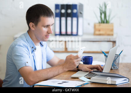 Portrait of happy young business man working in home office interior in loft space holding smartphone and looking at screen Stock Photo