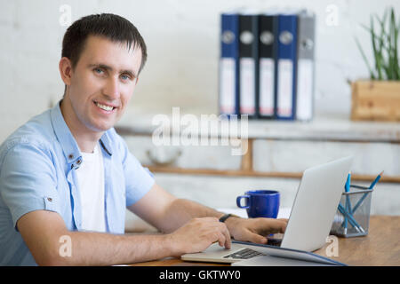 Portrait of cheerful young business man working on laptop in home office interior in loft space. Handsome guy sitting with lapto Stock Photo