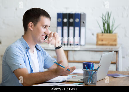 Portrait of cheerful young business man talking on phone in home office interior in loft space. Handsome guy sitting with laptop Stock Photo