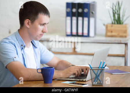 Portrait of young business man working on laptop in home office interior in loft space. Handsome serious guy sitting with laptop Stock Photo