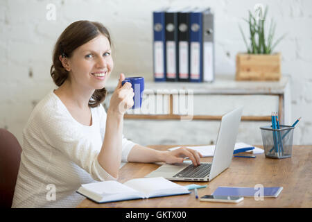 Portrait of young business woman drinking coffee and using laptop in home office interior. Happy casual office person sitting Stock Photo