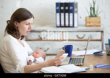 Portrait of young business mom holding her newborn cute babe while working in home office interior, looking at cellphone screen Stock Photo