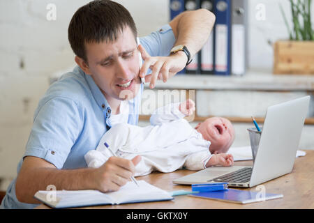 Portrait of young stressed dad trying to work and talk on phone while sitting with his newborn babe in home office interior. Stock Photo