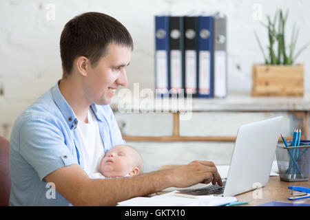 Side view portrait of happy casual young business dad holding newborn cute babe while working on laptop in home office interior. Stock Photo