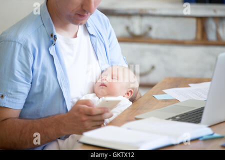 Casual business dad holding sleeping newborn babe while working in home office interior, holding smartphone and looking at scree Stock Photo