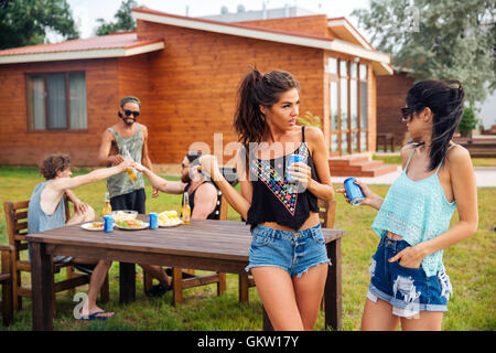 Two beautiful young women talking and drining soda with friends on summer outdoor party Stock Photo