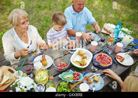 Family sitting at the table and eating grilled meat with vegetables outdoors Stock Photo