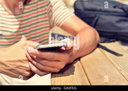 closeup of a young caucasian man sitting at an outdoor wooden table using his smartphone, with his backpack in the background Stock Photo