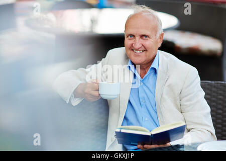 Happy senior man with open book and cup of coffee looking at camera in cafe Stock Photo