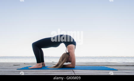 Beautiful sporty fit blond young woman in black sportswear working out outdoors on wooden pontoon on the lake, doing Bridge Pose, standing in Urdhva Dhanurasana, Chakrasana posture, full length Stock Photo