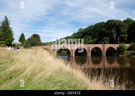 Bredwardine, Herefordshire, UK - A family picnic on the riverbank in summer sunshine on the River Wye as it passes under Bredwardine bridge. The brick bridge was built in the 1760s. Stock Photo