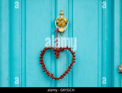 A heart shaped wreath hanging from a brass knocker on a turquoise coloured door Stock Photo
