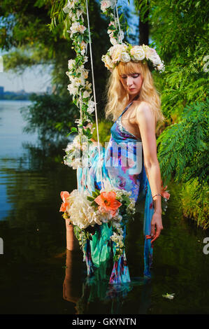 Girl on home made tree swing over river. Swing decorated with flowers. Woman with flower wreath on head. Stock Photo