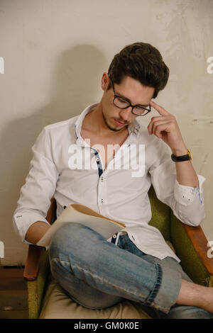 young man sits on the arm-chair holding his copybook on knee and thinks about something. There is his shadow on the wall behind Stock Photo