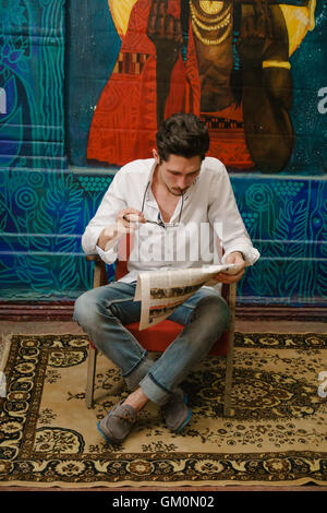 young man sits on the armchair reads a newspaper and bites his glasses. He noticed something interesting in this newspaper. Stock Photo
