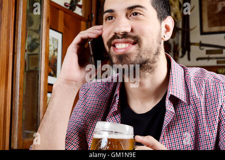 Portrait of young latin man talking on the phone with glass of beer and snacks in a bar. Indoors. Stock Photo