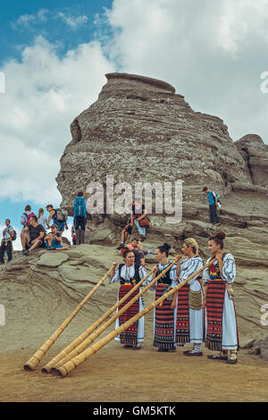 Bucegi Mountains, Romania - August 6, 2016: Young Romanian female artists wearing traditional costumes play the tulnic near the Sphinx megalith. Stock Photo