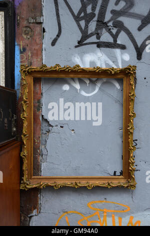 Blank vintage golden frame from antiques shop on cracked city wall with messy graffiti. Urban background. Stock Photo