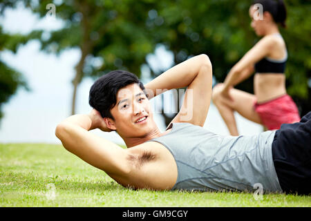 young asian man doing sit-ups on grass in city park. Stock Photo