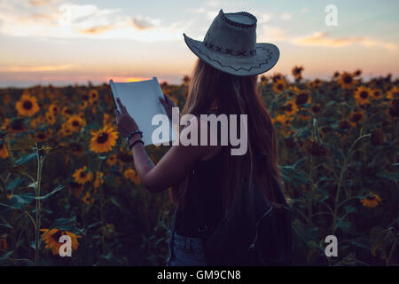 Girl in a cowboy hat in a sunflower field with a map looking for the path. Sunset Stock Photo
