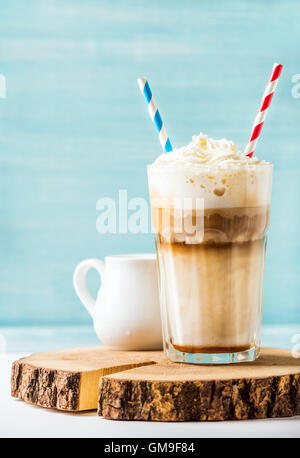 Latte macchiato with whipped cream in tall glass , two straws and pitcher on wooden board over blue painted wall background, copy space Stock Photo
