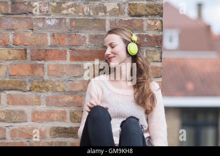Mid-adult woman with eyes closed and headphones on sitting by brick wall Stock Photo