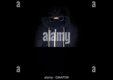 Cyber criminal in a hooded type with an unknown face sits at a laptop computer in the dark committing cyber crimes and hacking a Stock Photo